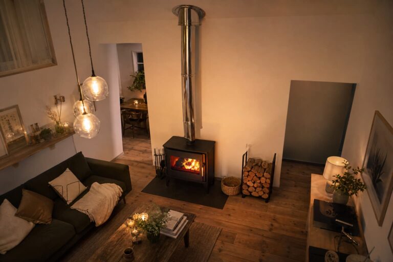 Une pièce à vivre cosy vue du dessus avec un poêle à bois moderne, un conduit de cheminée en acier inoxydable traversant un mur et un plafond blancs, une pile de bois de chauffage à proximité et un plancher en bois rustique, sous une lumière ambiante chaude.